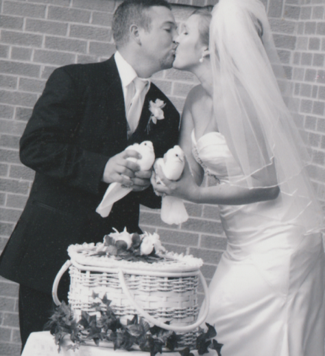 A married couple standing behind a white heart shaped basket holding two white birds.